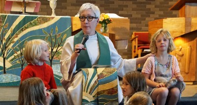 Female pastor, white robe, green stole, seated at front of sanctuary surrounded by children