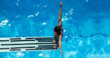 Man standing on edge of diving board over blue water in pool, with back to water and arms outstretched.