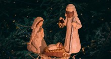 Wood carved figures of Mary, Joseph, Jesus in manger in front of Christmas tree with small lights.