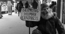 black and white photo of a man with white bushy beard in a Boston subway station holding a sign that says "seeking human kindness"